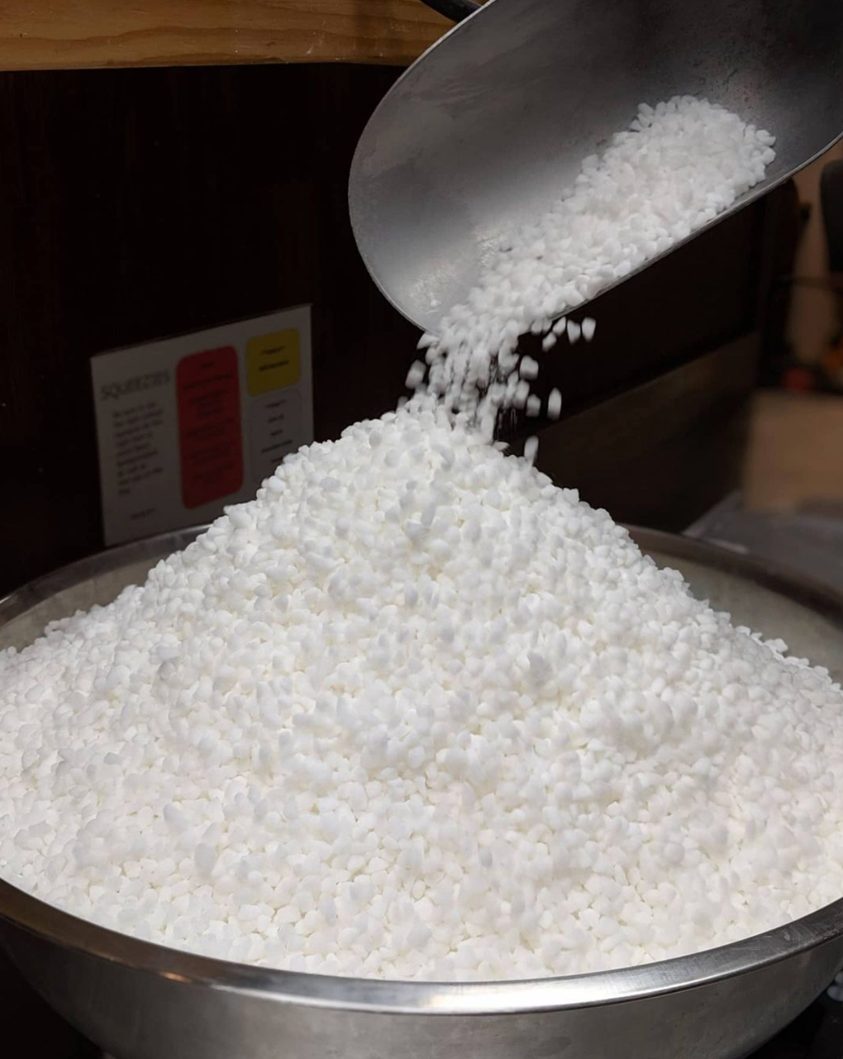 A metal scoop pouring "Sugar Pearls" into a large metal bowl already filled with the same pellets.