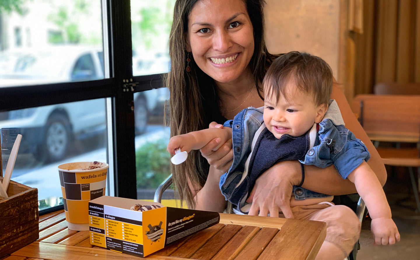 Happy mother holding her toddler who’s trying to reach a chocolate waffle with his spoon