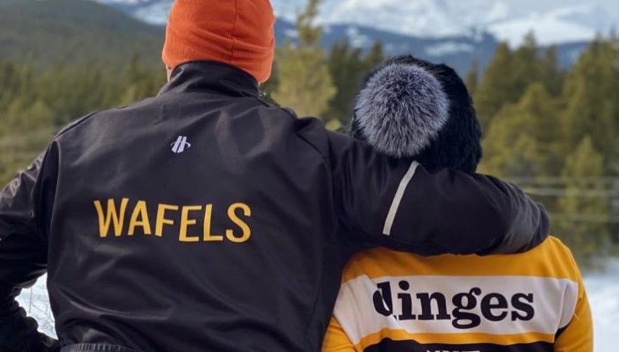 A couple in the winter cold,  one jacket reads "wafels"  and the other "dinges."